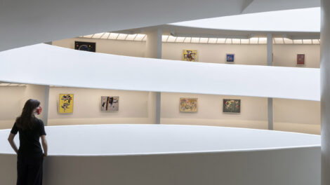 A person standing with their back to the camera and looking across the Solomon R. Guggenheim's rotunda to see colorful abstract paintings hanging along the spiraling museum walls.