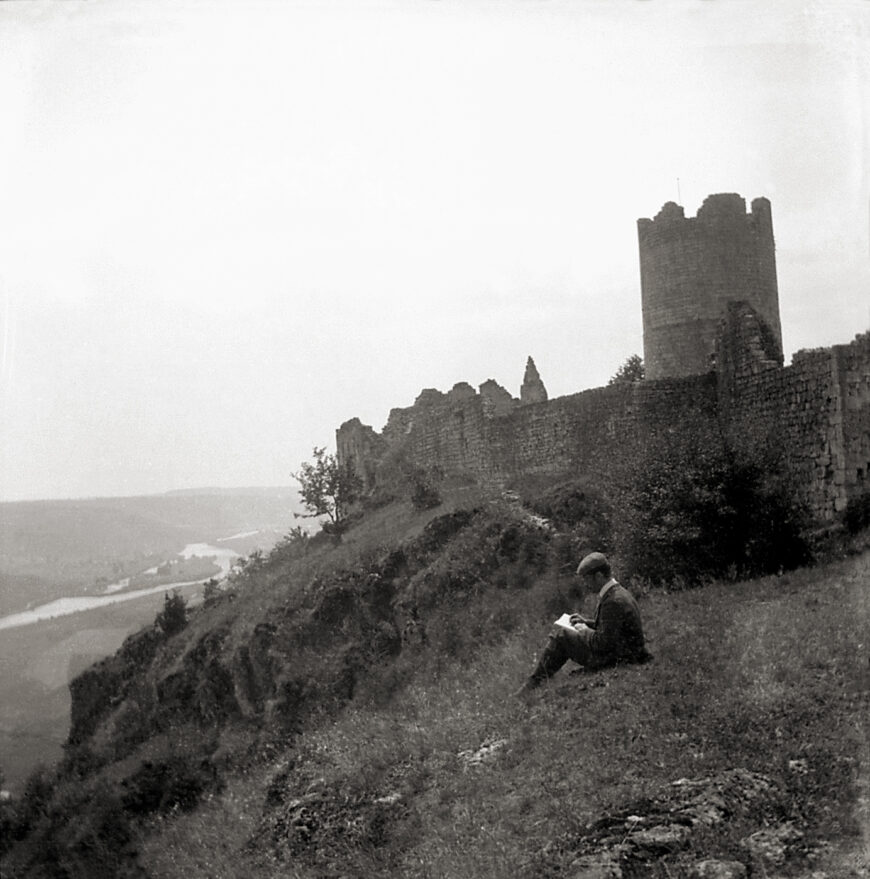 Black and white photograph of a man on a grassy hill looking at a sketchbook