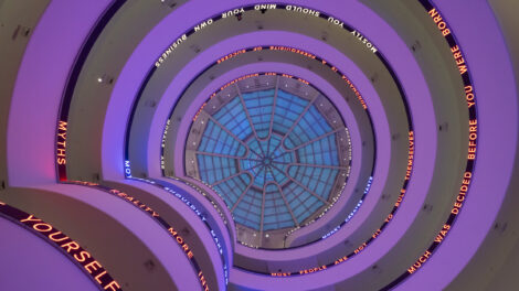 Walls of the Guggenheim Museum bathed in a purple glow. A scrolling LED text installation winds up the ramps of the Guggenheim Museum, displaying texts written and curated by the artist, leading to a blue sky beyond the oculus.